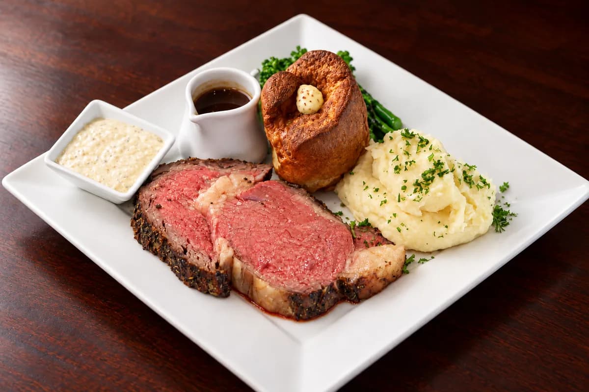 Sliced prime rib with a dark roast crust, Yorkshire pudding with butter, broccolini, garlic mashed potatoes, au jus and creamy horseradish on a white plate.