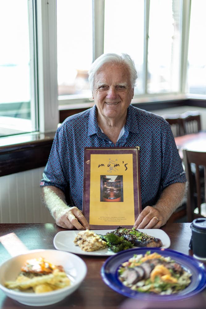 John Morris seated at the window at Boathouse on the Bay, holding the original Mum's menu, with the three returning dishes plated in front of him